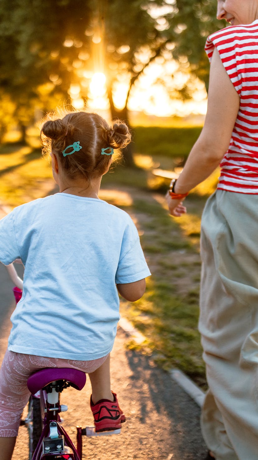 A little girl learning to ride a bike in the park, with the help of her mother. Rear view photo.