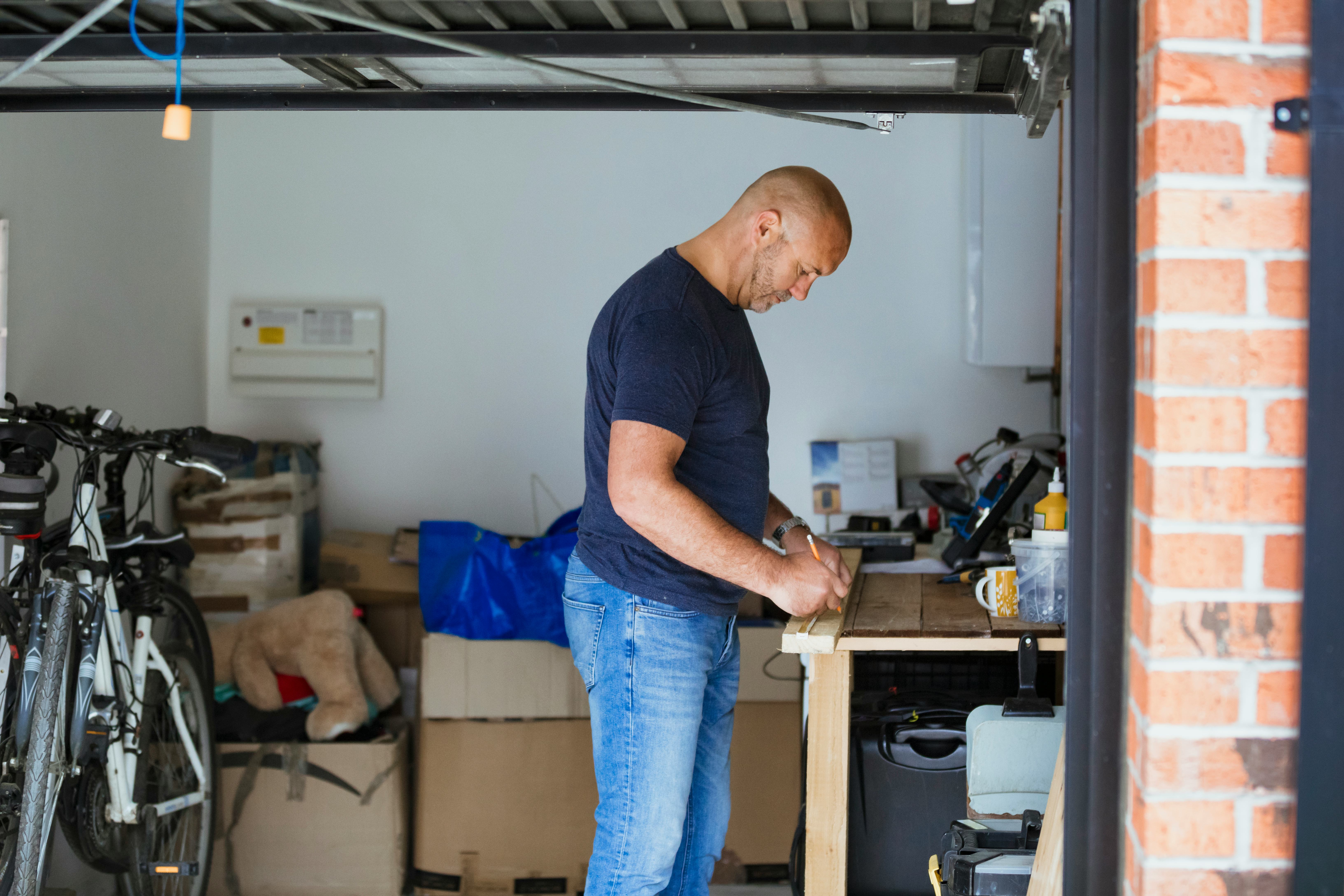 A side-view shot of a male carpenter upcycling, he is measuring a plank of wood on a crafting table, he is wearing casual clothing.