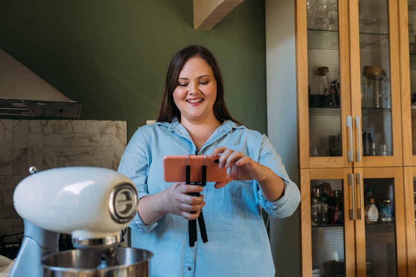 A happy plus size professional female chef setting up her mobile phone to film herself cooking in a …