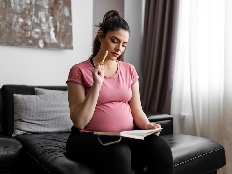 Pregnant woman sitting on sofa at home and taking notes. Mother-to-be. Expecting a new life concept