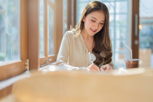 Smiling portrait of asian freelance female adult remote working writing a novel book in cafe morning...