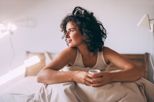 A content woman with curly hair enjoys a peaceful morning, sipping coffee in bed. The tranquil atmos...