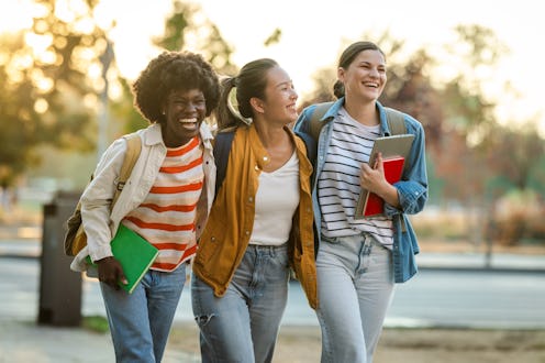 Group of three diverse young women walking and smiling outdoors in a city setting, enjoying sunny we...