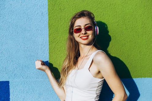 Happy young woman in red sunglasses and white top listening music while standing close to squared co...