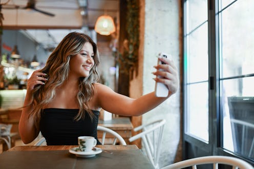 Beautiful Smiling Female Adult Checking Hair on Her Smartphone While Drinking Coffee