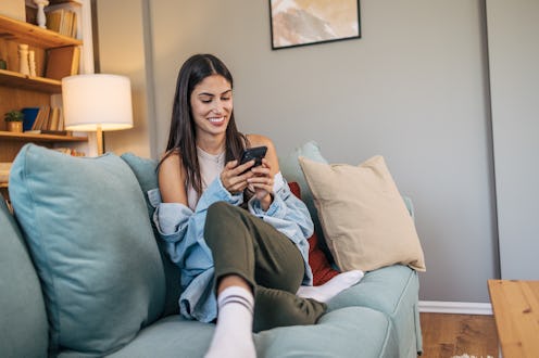 A young cheerful woman relax on sofa and using smartphone.
