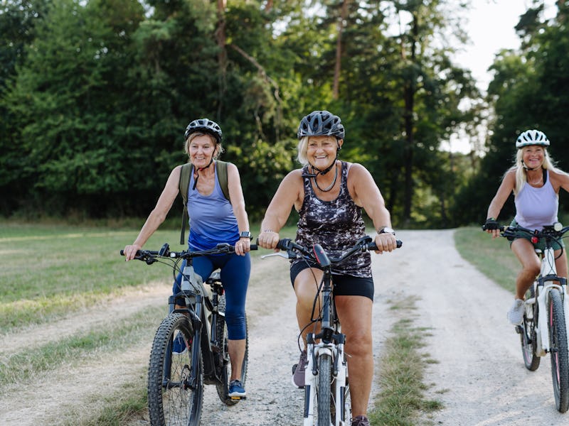 Three senior friends on bike tour in nature enjoying ride. Older women on low impact route riding el...