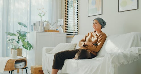 Relaxed Asian woman sick with cancer with a headscarf enjoys her leisure time watching television, s...