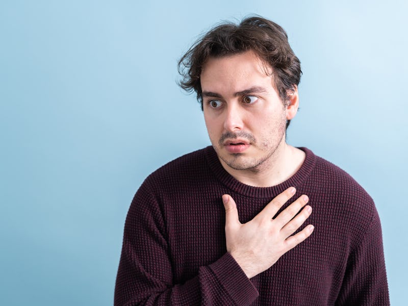 A young man with chest pain on a blue background. He looks disconsolately to the side and has his ha...
