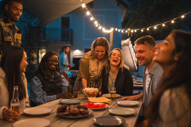 A joyful gathering of diverse male and female friends laughing and dining at an outdoor party, with ...