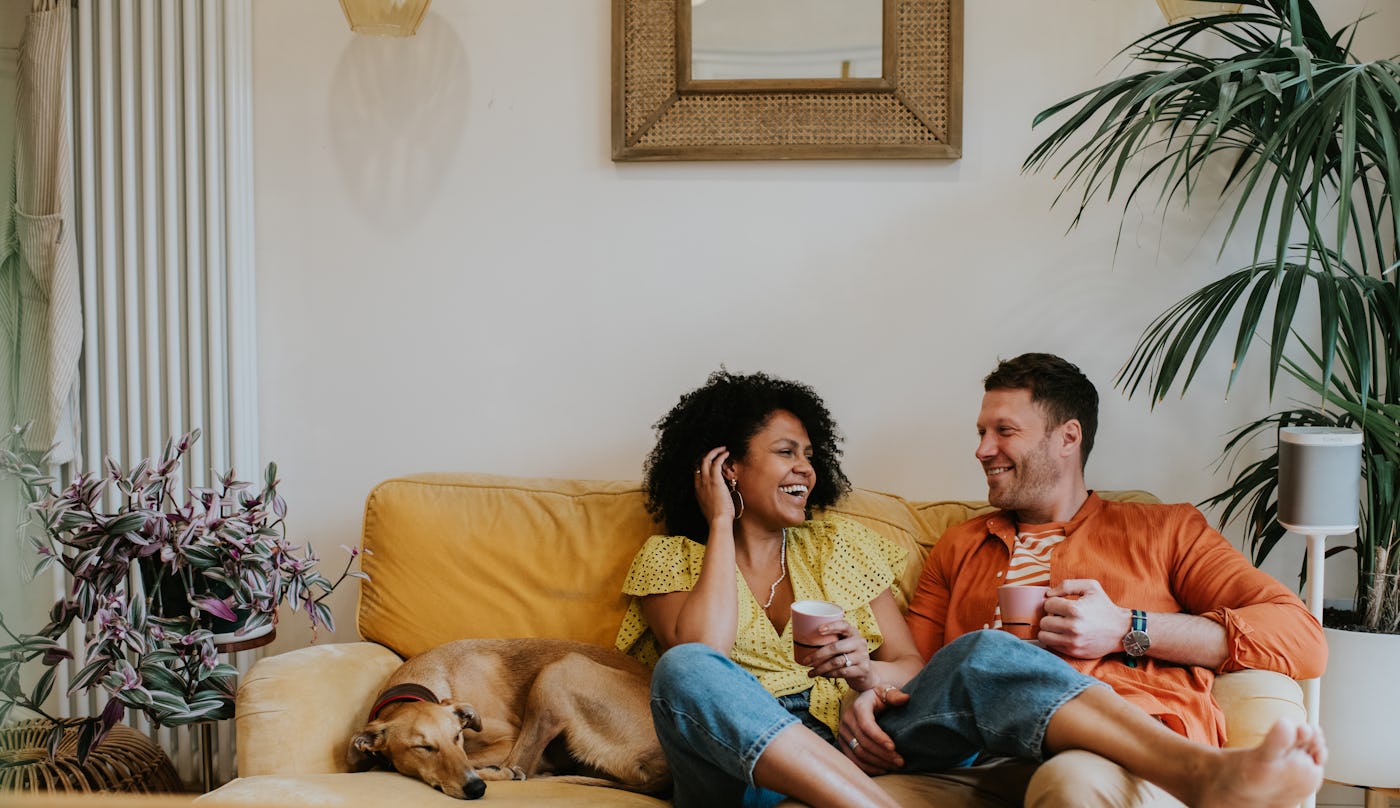 A beautiful black woman sits with her leg draped over her male partners thigh. They are comfortable and happy in a stylish living room on a soft, velvet, yellow sofa. They giggle. A lurcher dog is curled up and naps beside them. Long wall mounted modern radiator visible in the background Space for copy.