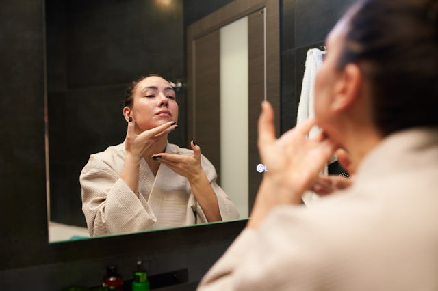 Woman in cotton bathrobe applying cream and massaging chin while looking at mirror during beauty rou...