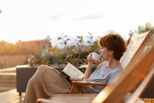 Beautiful woman is sitting on patio on the deck chair and enjoying beautiful surrounding while drink...
