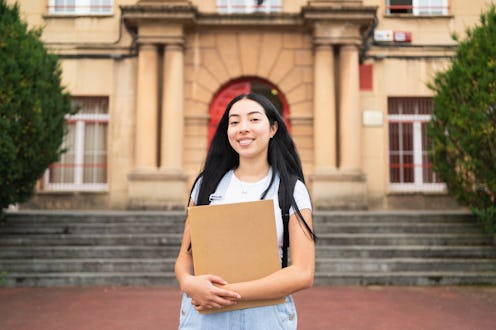 portrait of a young hispanic woman is smiling while holding a book, she is standing in front of her ...