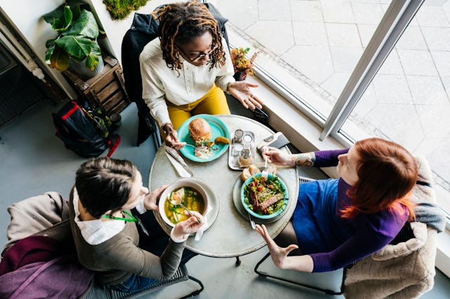 An aerial view of three women sitting at a small table in a vegan cafe, eating colorful plates of fo...
