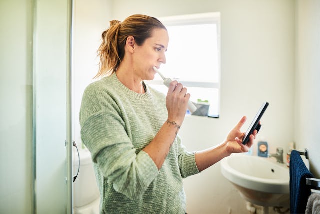 Woman standing in her bathroom at home brushing her teeth with an electric toothbrush and checking h...
