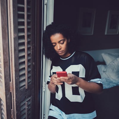 Depressed teenager with curly hair wearing a sports jersey texting as they lean against a window frame.