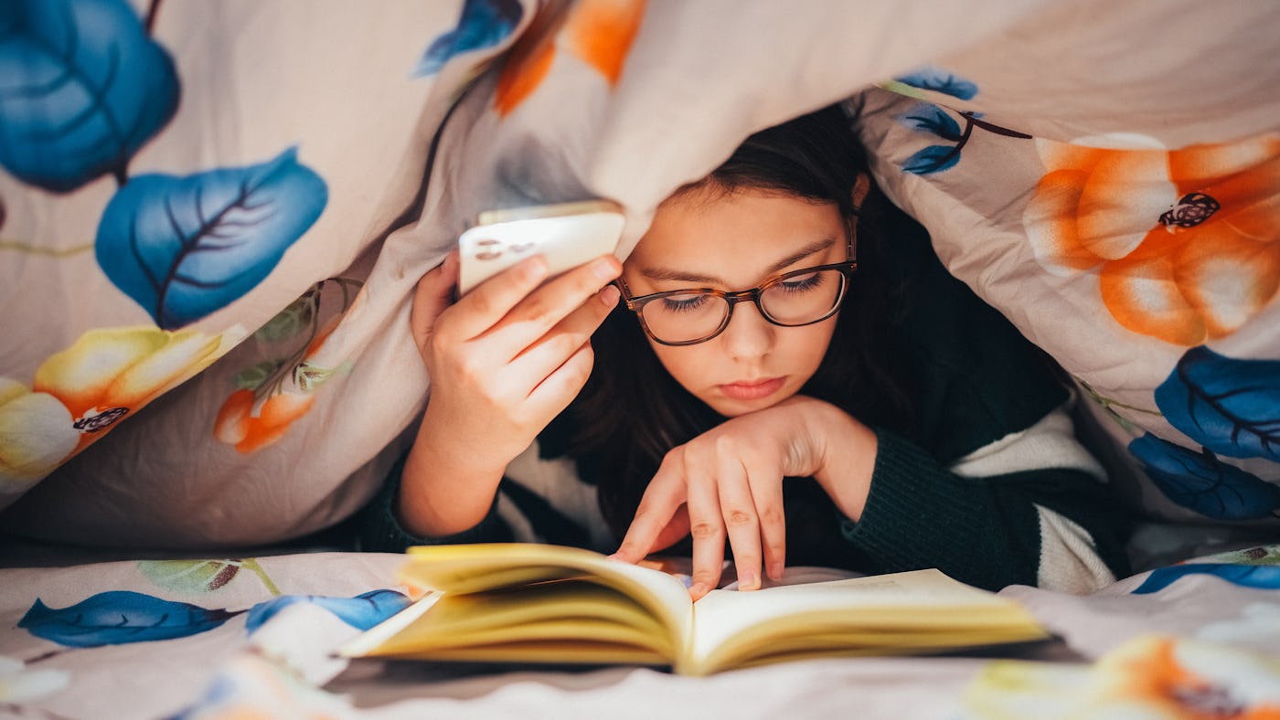 Girl lights with phone lantern under blanket to read a book late at night