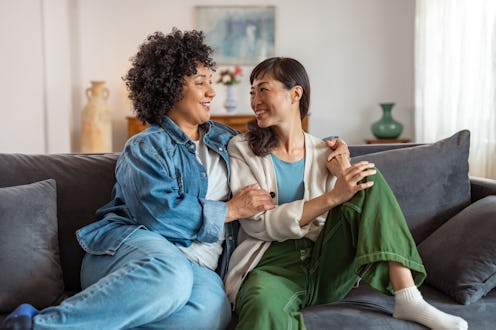 A loving diverse lesbian couple sits comfortably on a couch, sharing a tender moment at home. The at...