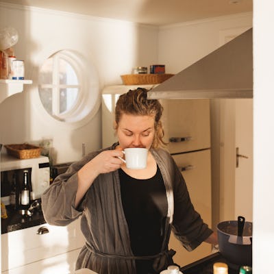 A woman in a cozy kitchen, wearing a robe, enjoys a cup of coffee while standing near a stove. Natural light brightens the space.