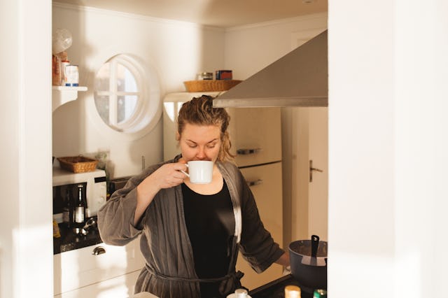 A woman in a cozy kitchen, wearing a robe, enjoys a cup of coffee while standing near a stove. Natur...
