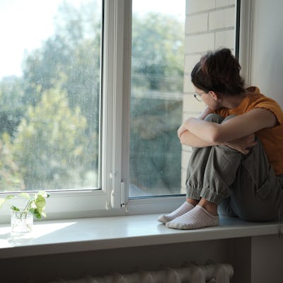 Sad upset teenager sitting on windowsill hugging knees looking out window, lonely teen girl at home. Adolescent Loneliness
