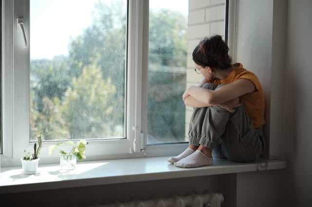 Sad upset teenager sitting on windowsill hugging knees looking out window, lonely teen girl at home....