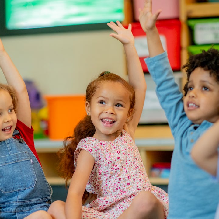 A small group of preschool children are sitting on the floor of their classroom with their hands rai...