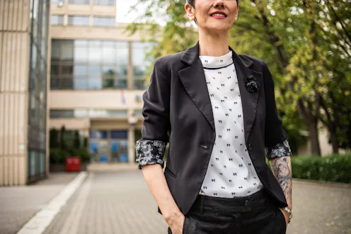A mature woman with short hair, dressed elegantly, stands with crossed arms with a tattoo on them