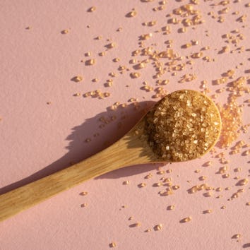 Brown cane sugar in spoon on pink background in bright sunlight