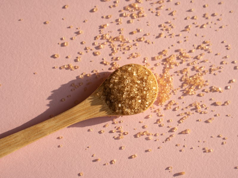 Brown cane sugar in spoon on pink background in bright sunlight