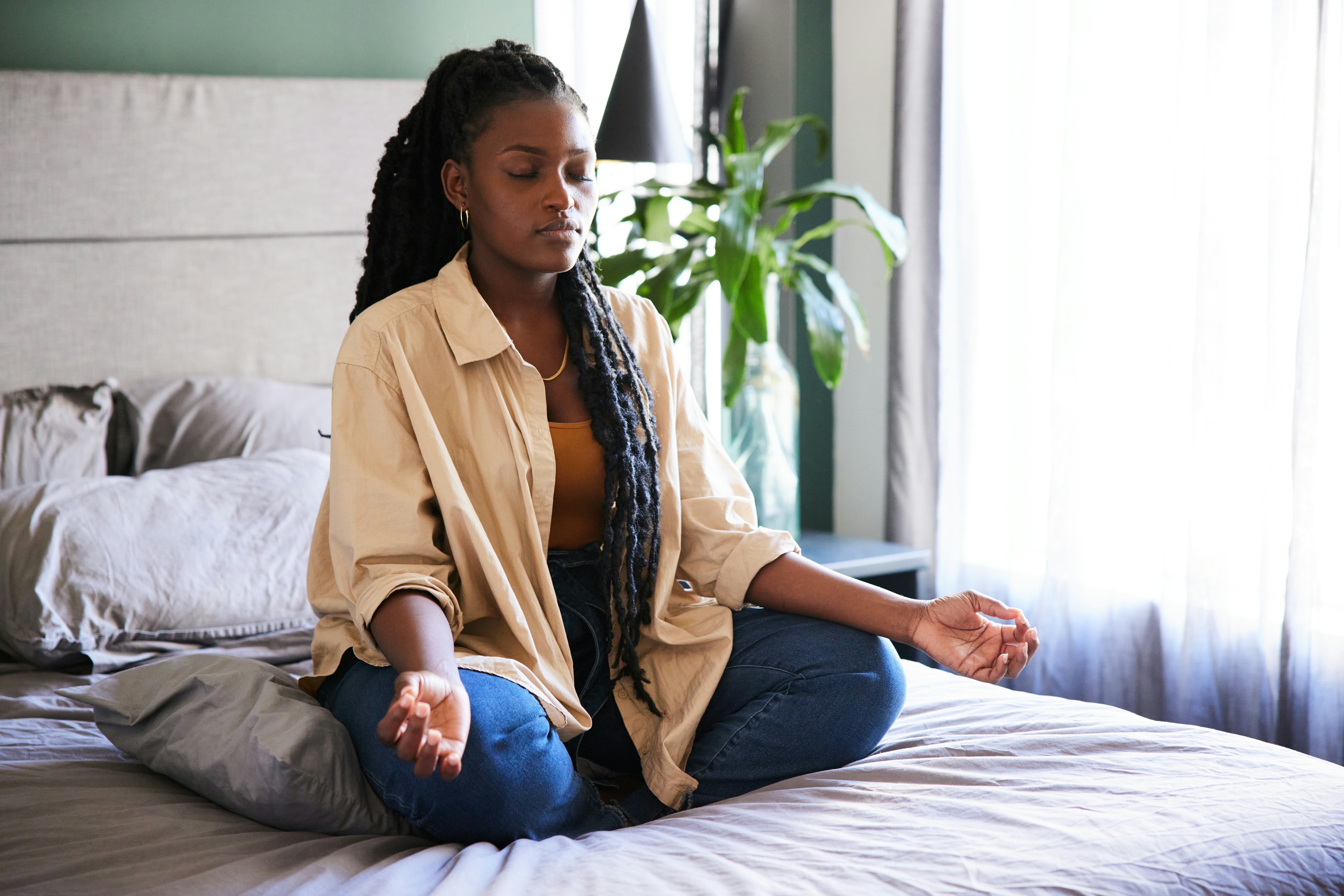 Young African woman meditating with her eyes closed in the lotus position on her bed at home in the &hellip;