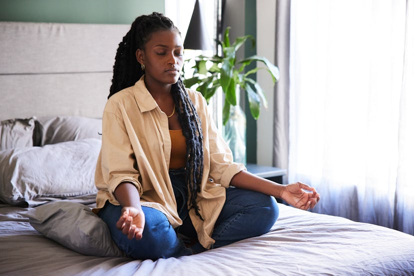 Young African woman meditating with her eyes closed in the lotus position on her bed at home in the …