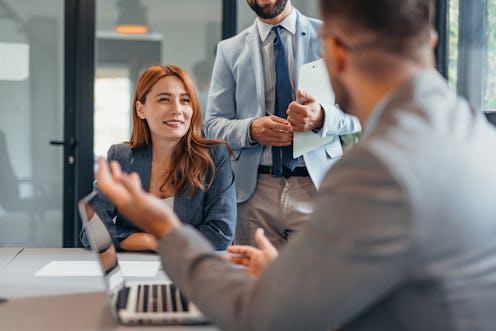 In a professional office setting, three colleagues engage in a productive meeting. A woman smiles wh...