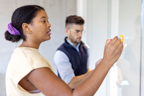 Businesswoman working on adhesive note over glass wall with male colleague in background. Young woma...