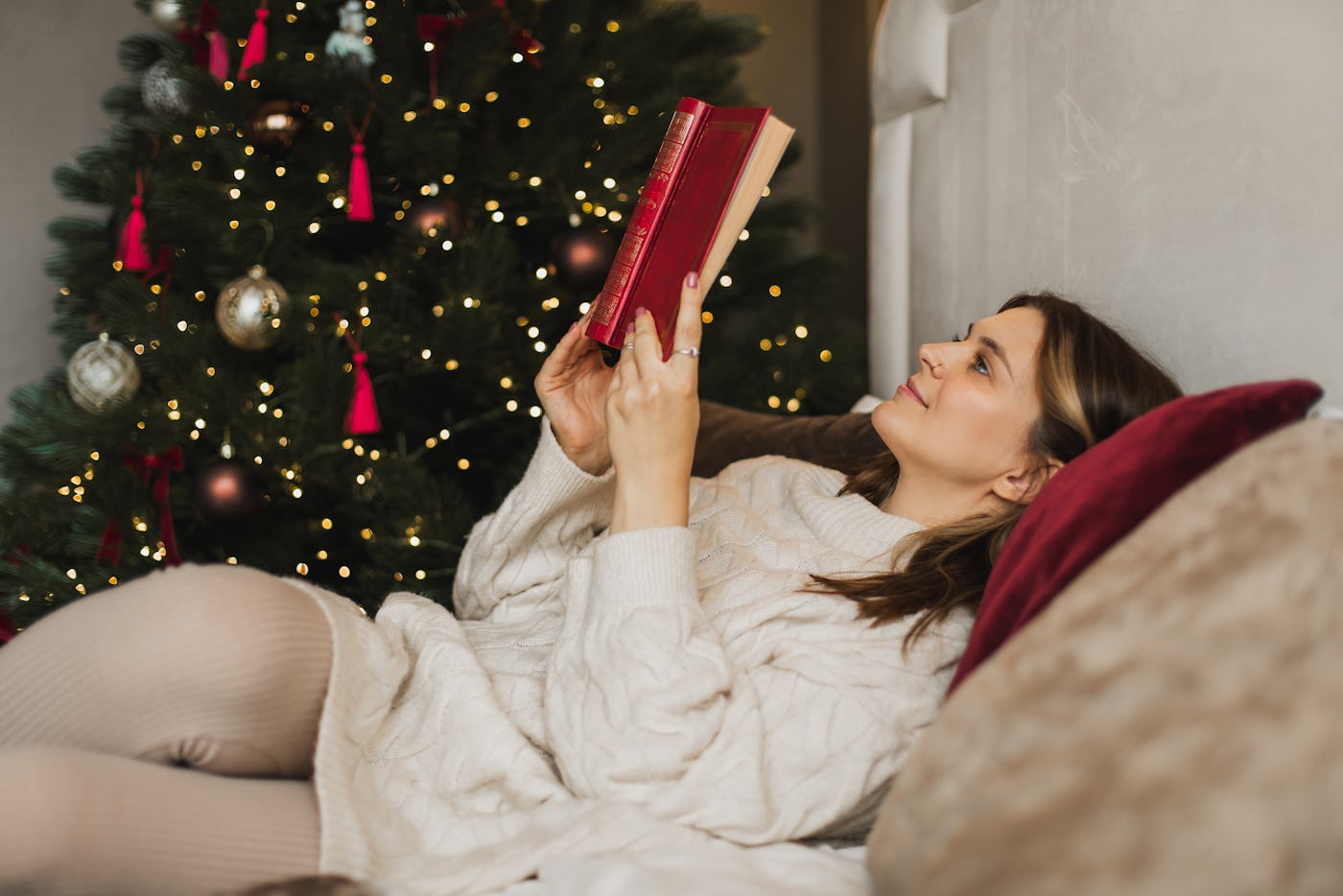 Pretty cute woman in wool sweater is reading book on the bed near Christmas tree. Relaxing and hobbies during holidays. Christmas and holidays concept.