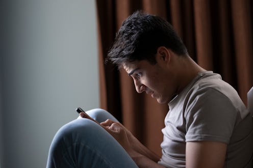 Young man is sitting on his bed, looking at his smartphone with a shocked and disbelieving expressio...