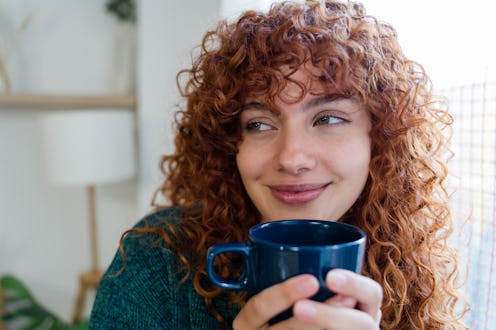 Young redhead woman with curly hair smiling and holding a blue mug at home, enjoying a moment of rel...