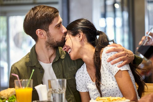 Front view of young adult man sitting at mall's food court table while kissing girlfriend's fronthea...