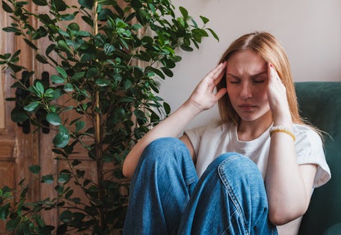 Young unhappy depressed woman suffering from headache, sitting in armchair holding head in hands fee...