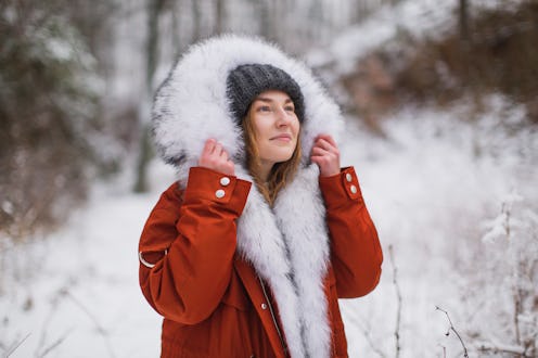 Redhead woman in red jacket with fur and hat in winter forest. Pretty woman enjoying vacation. Girl ...