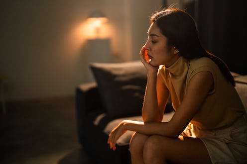 A thoughtful woman sits on a sofa, illuminated by gentle evening light, conveying introspection and ...