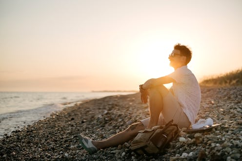 Handsome young man sitting thoughtful on the beach looking in the far