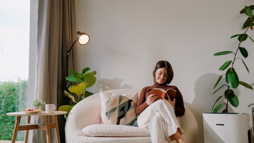 A young Asian woman turning on an air purifier in the living room before sitting on an armchair and ...