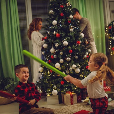 Candid shot of two little kids having fun playing with wrapping paper rolls they are using as swords while their parents are decorating the Christmas tree in the background.