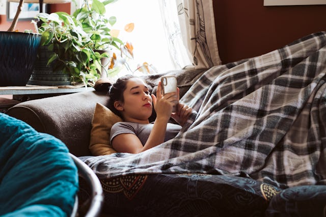 Teenage child lying on couch under a blanket watching something on phone.