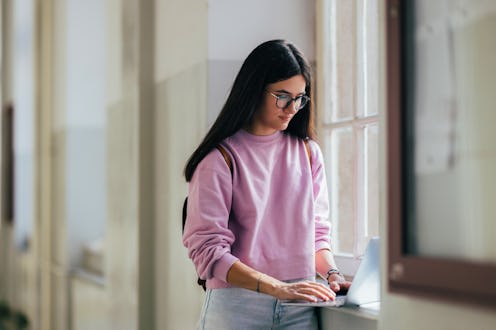 A young woman in a pink sweatshirt works on a laptop by a window, reflecting concentration and focus...