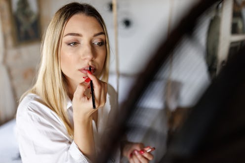 Close-up shot of beautiful young woman standing in front of a mirror in bedroom and putting lipstick...