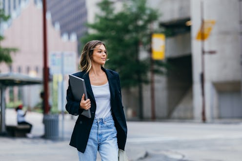 A confident young businesswoman in casual attire holding a laptop while walking in an urban setting....