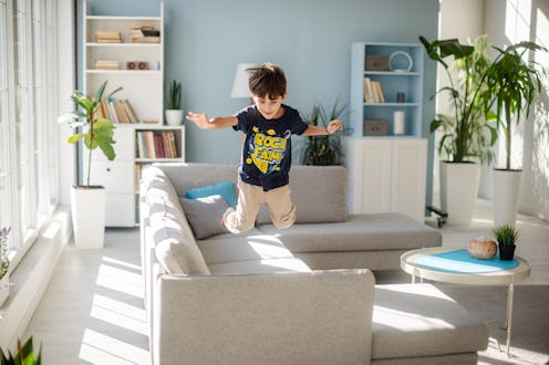 A dynamic scene of a young boy energetically jumping and leaping on a soft grey couch in a stylish, ...